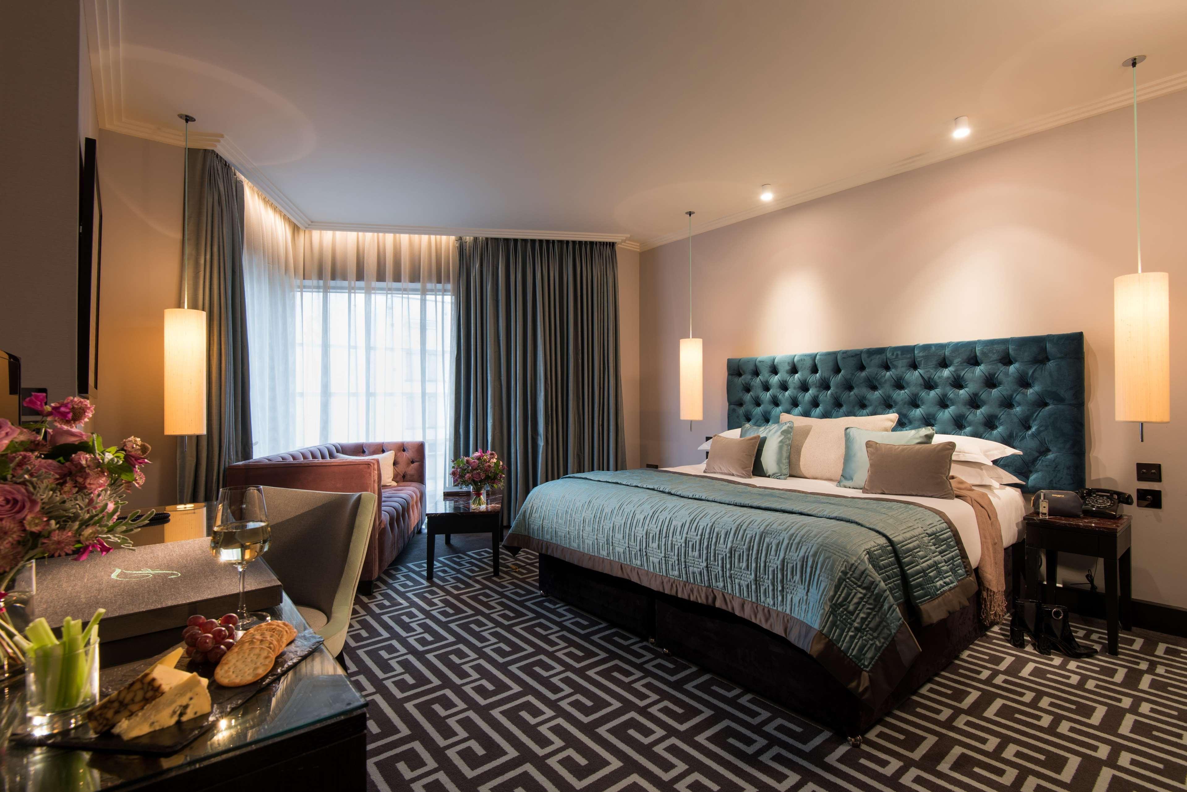 The Fitzwilliam Hotel Belfast Exterior foto The photo depicts a stylish hotel room. It features a large bed with a tufted teal headboard, flanked by two wall sconces providing soft lighting. The bedding is neatly arranged with accent pillows. To one side, there is a small seating area with a c